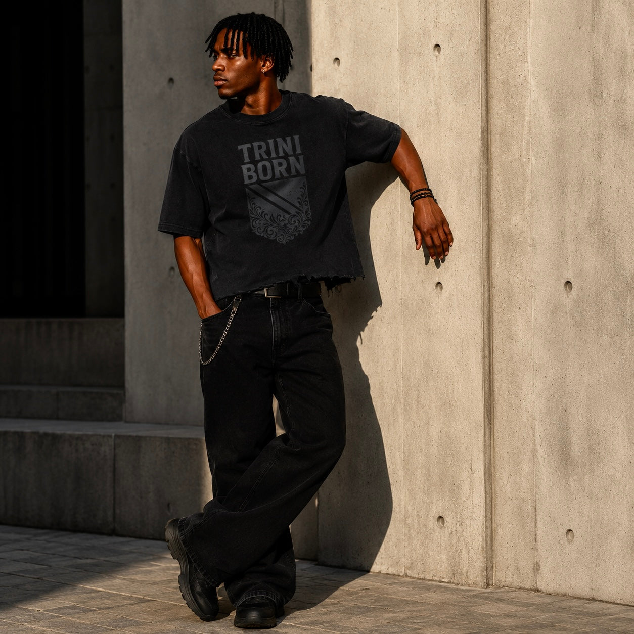 Black male model wearing the Trini Born Culture Tee in vintage black with a gray distressed TRINI BORN crest graphic, styled with oversized black jeans and black sneakers while leaning against a concrete wall in warm natural light. The oversized boxy T-shirt features a cropped distressed hem that sits at the belt line, creating a modern luxury streetwear look.