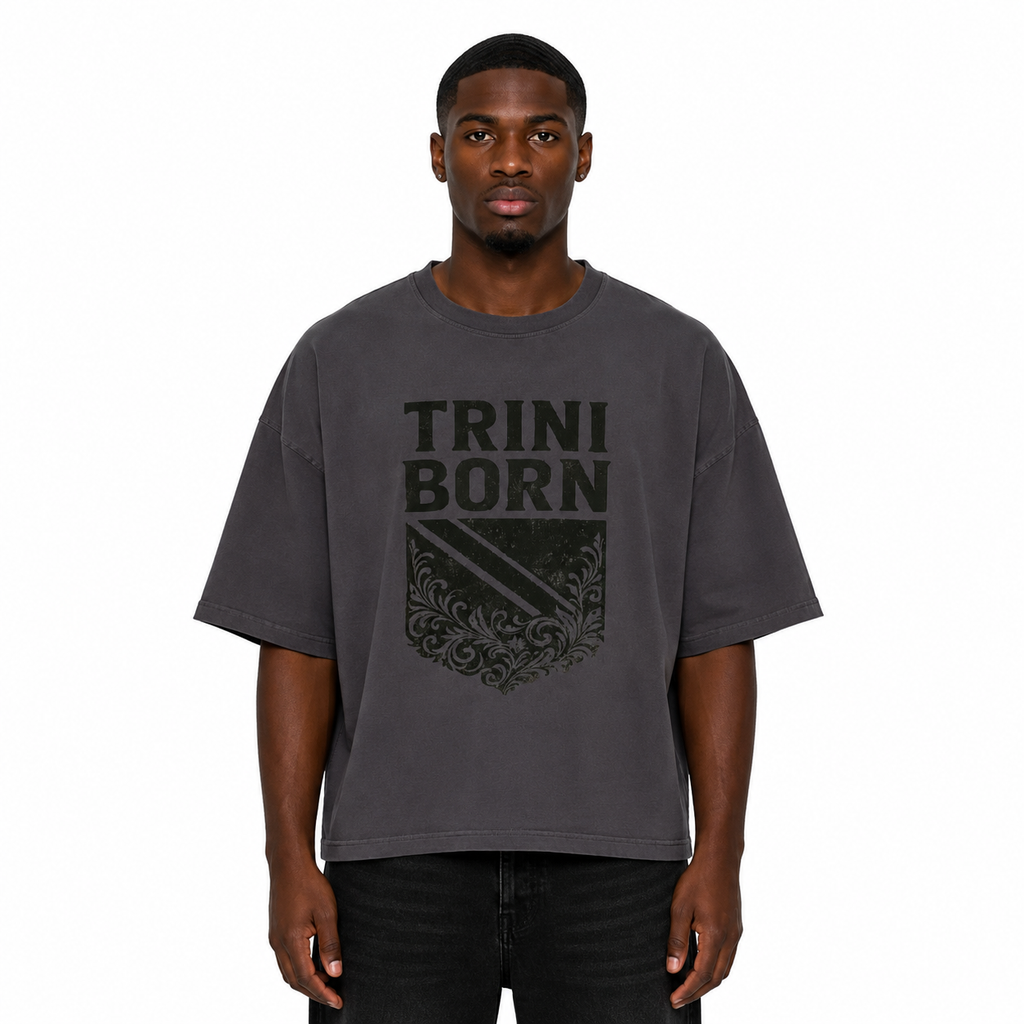 Black male model wearing the Trini Born oversized charcoal graphic T-shirt with black crest artwork, relaxed boxy fit, front view on a white studio background.
