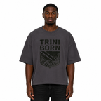 Black male model wearing the Trini Born oversized charcoal graphic T-shirt with black crest artwork, relaxed boxy fit, front view on a white studio background.