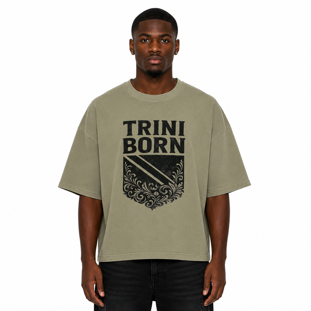 Black male model wearing the Trini Born oversized Gray Green graphic T-shirt with black crest design, relaxed boxy silhouette, front view on a white studio background.
