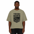 Black male model wearing the Trini Born oversized Gray Green graphic T-shirt with black crest design, relaxed boxy silhouette, front view on a white studio background.