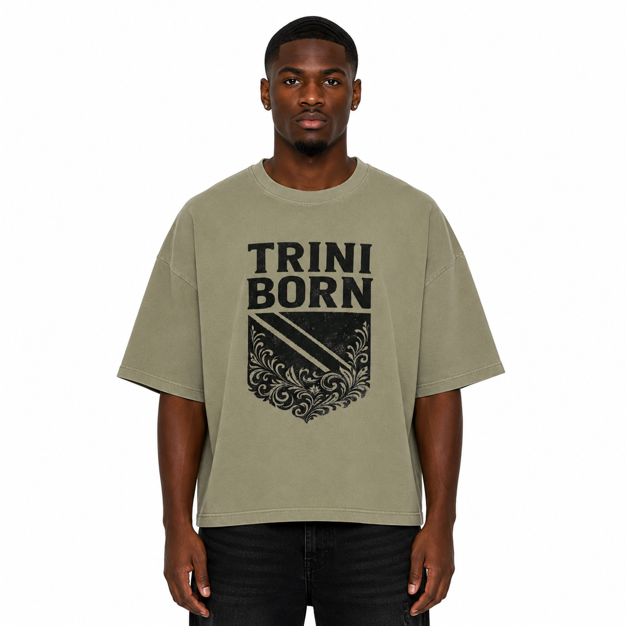 Black male model wearing the Trini Born oversized Gray Green graphic T-shirt with black crest design, relaxed boxy silhouette, front view on a white studio background.