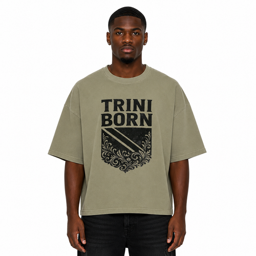 Black male model wearing the Trini Born oversized Gray Green graphic T-shirt with black crest design, relaxed boxy silhouette, front view on a white studio background.