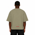 Back view of Black male model wearing the Trini Born oversized Gray Green T-shirt featuring a red Trini Born logo near the lower back on a white studio background.
