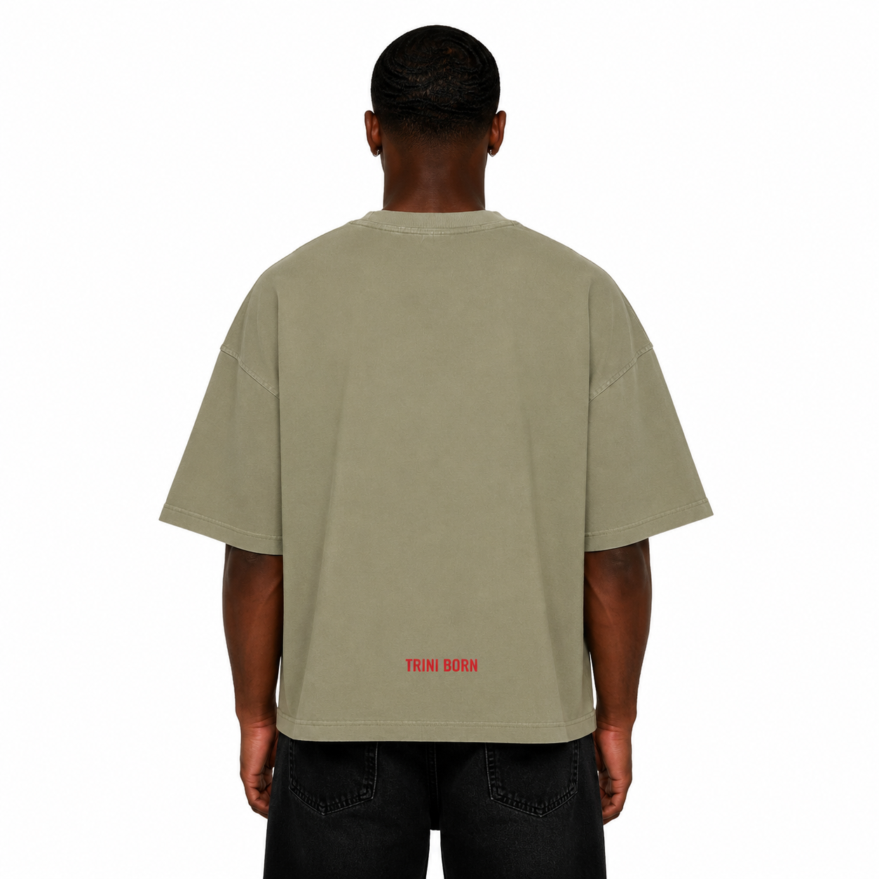 Back view of Black male model wearing the Trini Born oversized Gray Green T-shirt featuring a red Trini Born logo near the lower back on a white studio background.