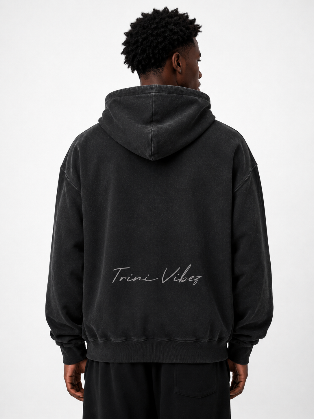 Back view of Black male model wearing the Trini Born Culture Raised Me heavyweight oversized hoodie in washed black with tonal Trini Vibez script.