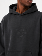 Close-up of the washed black Trini Born Culture Raised Me heavyweight oversized hoodie on a Black male model, showing the tonal THE CULTURE RAISED ME. chest design and sunfaded cotton finish.