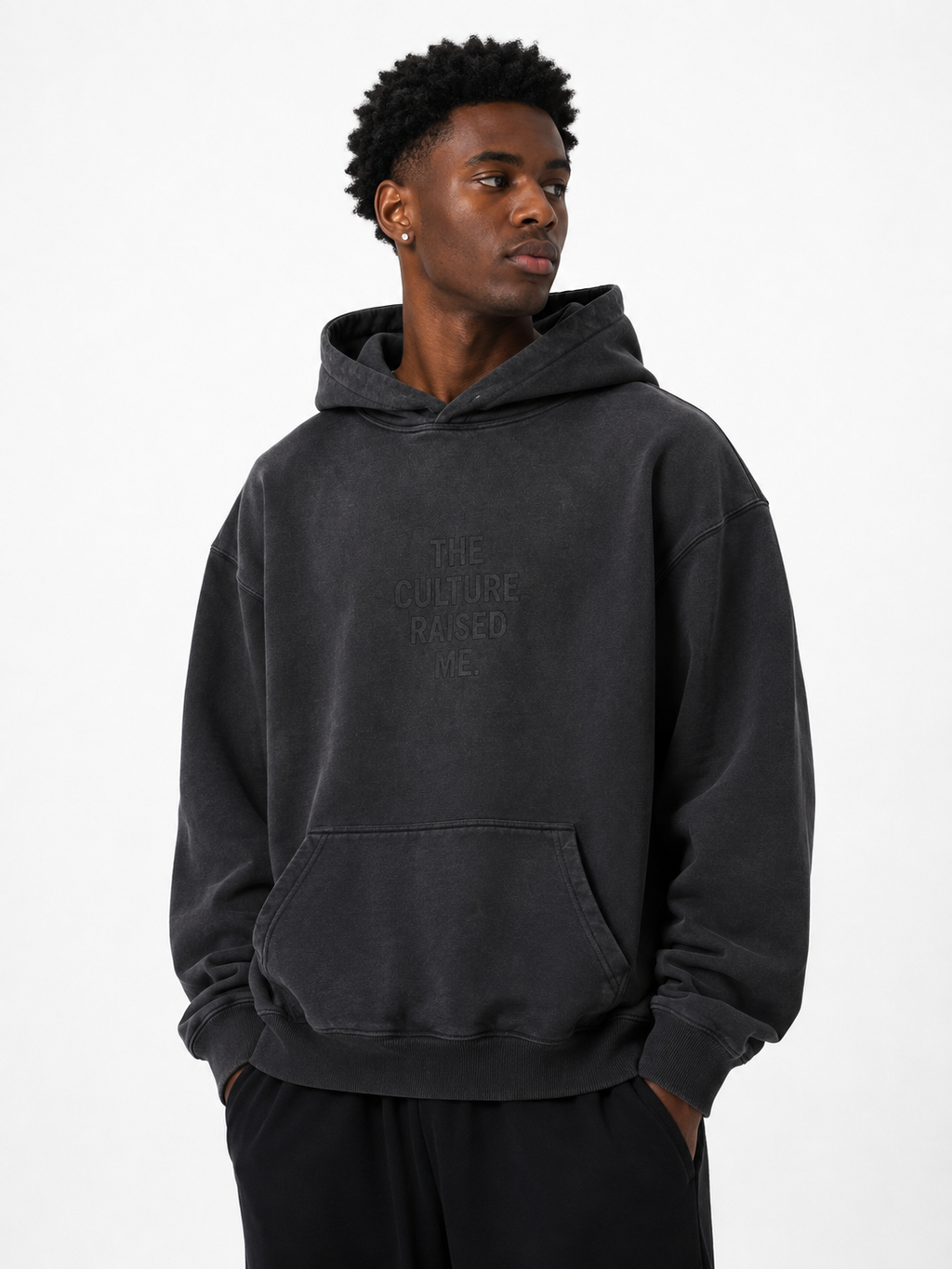 Black male model wearing the Trini Born Culture Raised Me heavyweight oversized hoodie in washed black, front view with tonal THE CULTURE RAISED ME. graphic.