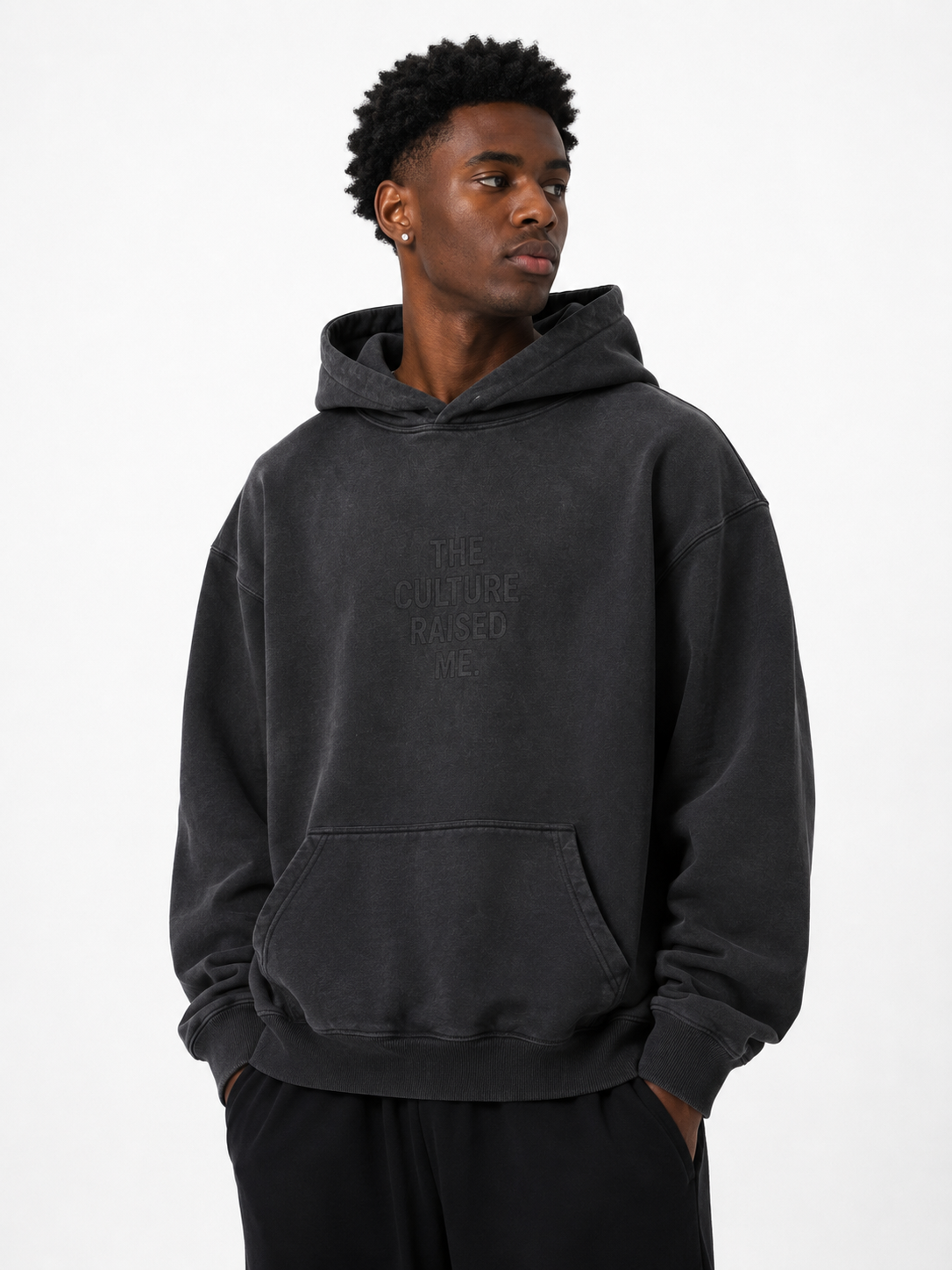 Black male model wearing the Trini Born Culture Raised Me heavyweight oversized hoodie in washed black, front view with tonal THE CULTURE RAISED ME. graphic.