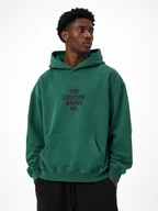 Black male model wearing the Trini Born Culture Raised Me heavyweight oversized hoodie in washed green, front view with tonal THE CULTURE RAISED ME. graphic.
