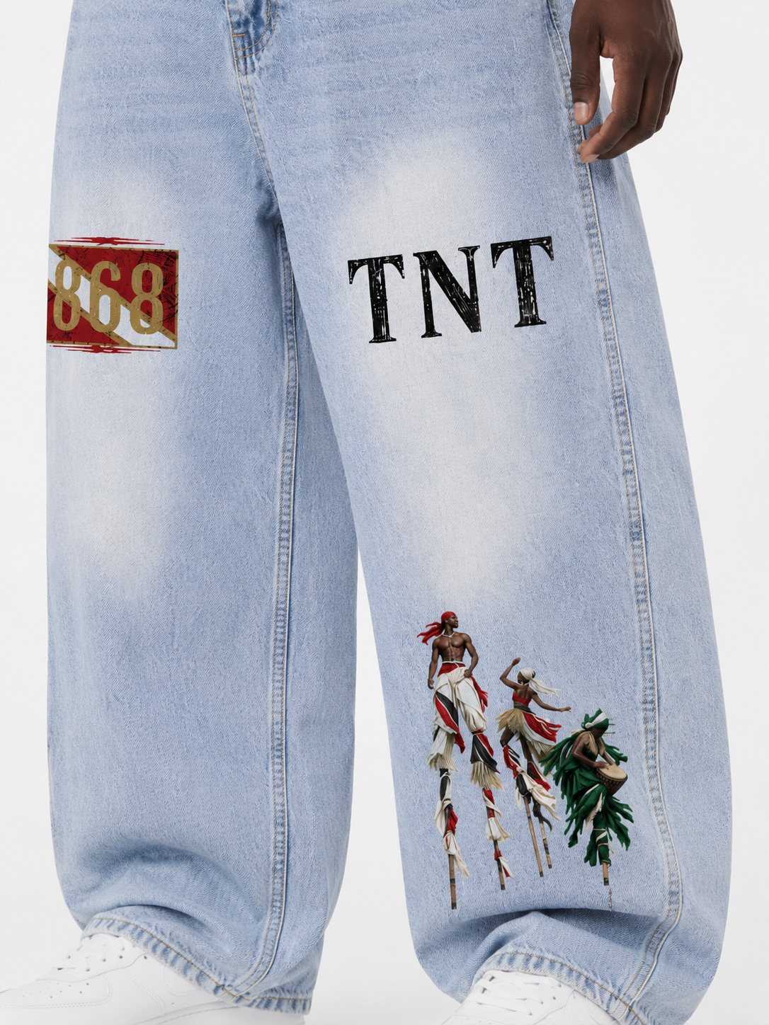 Close-up of Trini Born light blue vintage wash barrel leg jeans on a Black male model showing the 868, TNT, and Trinidad carnival stilt performer artwork.